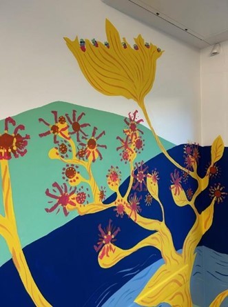 A close up of the mural in a corner showing a yellow plant with red flowers trailing up the wall.