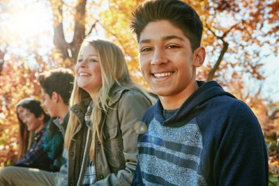 A teenage boy smiles at the camera. Three other teenagers sit behind him outside, not looking at the camera.