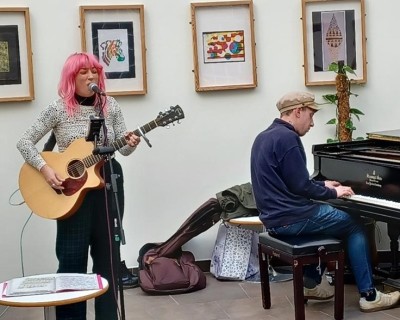 A woman is stood playing an acoustic guitar and singing into a microphone. A man is beside her playing the piano.