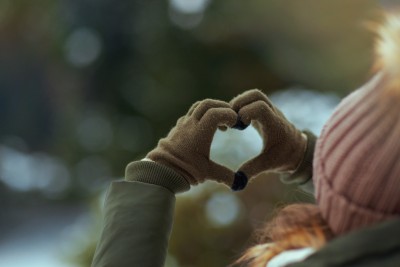 A woman outside dressed in a hat and scarf faces away from the camera. Using her hands, she makes a heart shape and holds it up into the sky.