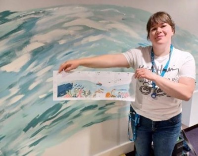 A woman holding a printed version of the mural design in front of the mural being painted
